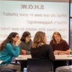 four students at table with projection on screen in background, asking SHOW, what do we see in your photo?